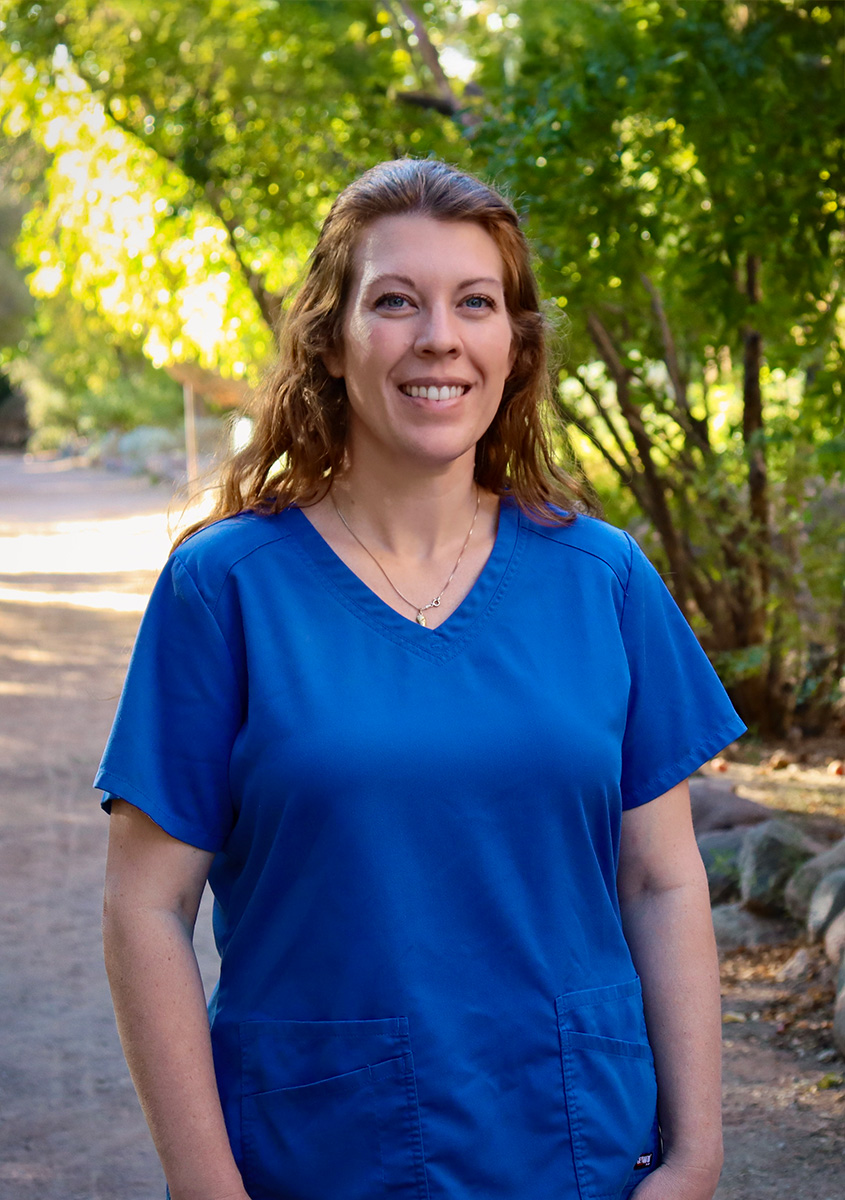 Smiling woman in a blue scrub top standing on a tree-lined path.